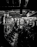 A dimly lit casino filled with rows of slot machines, some occupied by people. The machines' lights contrast with the dark surroundings, creating a vibrant yet subdued atmosphere.