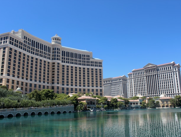 A large luxury hotel and casino building with multiple stories, featuring a light facade with arched windows. The structure has a prominent central tower with a dome-like top. It is surrounded by landscaping with trees and a body of water in the foreground.