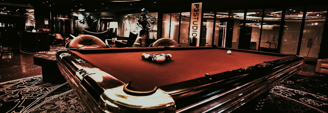 A luxurious interior features a billiard table with balls arranged on it. The ceiling has an intricate stained glass design, and the space is dimly lit, creating a sophisticated atmosphere. Comfortable chairs are placed around the area, with some decorative plants and a lamp adding to the elegance.
