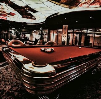 A luxurious interior features a billiard table with balls arranged on it. The ceiling has an intricate stained glass design, and the space is dimly lit, creating a sophisticated atmosphere. Comfortable chairs are placed around the area, with some decorative plants and a lamp adding to the elegance.